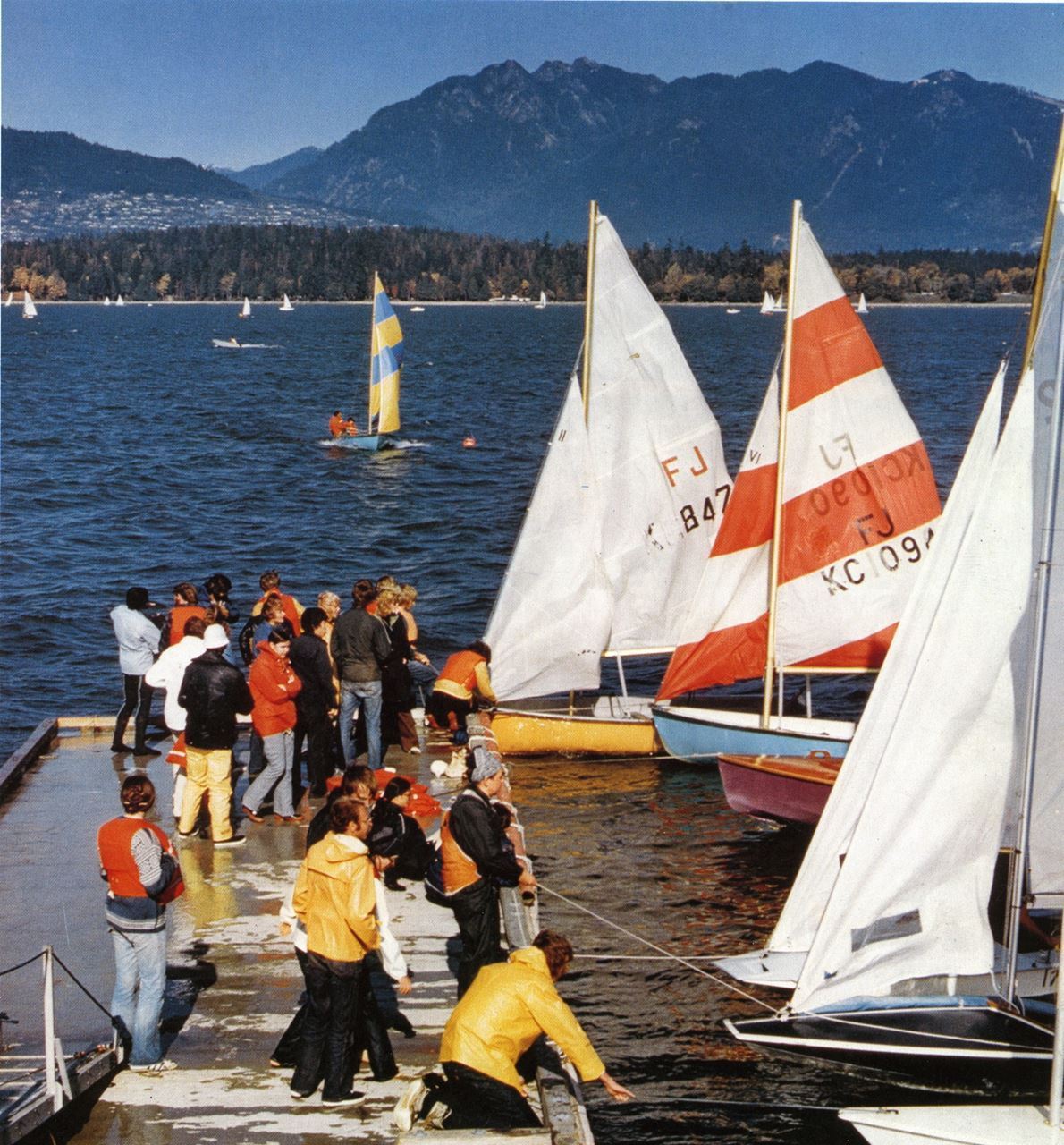 UBC Flying Juniors at the Kitsilano dock. The Fireballs in the foreground are KYC members’ boats. This picture appeared in the Winter issue of the Beautiful BC magazine. I’m the guy wearing the yellow oilskin jacket, standing in the foreground. A couple of KYC Fireballs are also visible at the lower right, as well as a sliver of a Laser. 